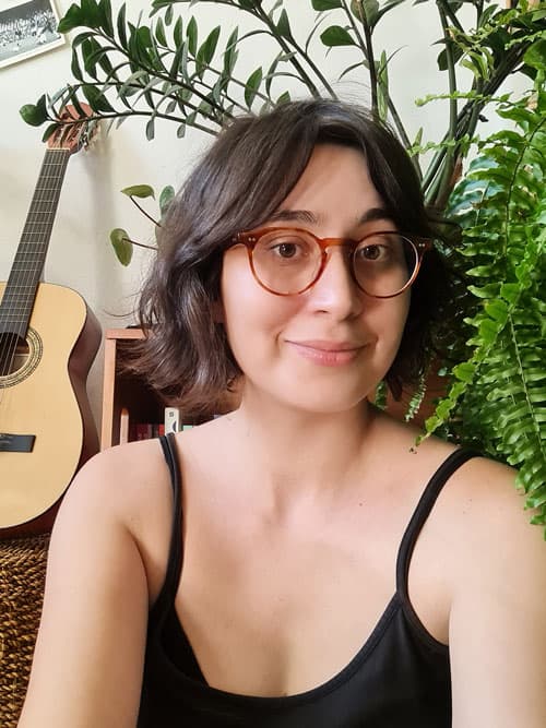 Ana Caroline é uma mulher branca de cabelos curtos, lisos e castanhos. Sorri levemente para a câmera. Usa uma camiseta preta e óculos de cor caramelo e, ao fundo, vê-se uma samambaia