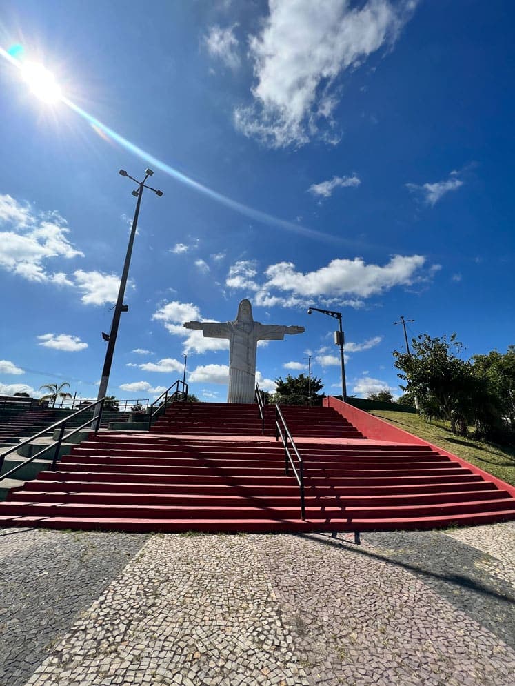 A imagem retrata a paisagem da Praça do Cristo, no Barreiro. Ao centro, sob o céu azul em um dia de tempo aberto, ganha destaque uma imagem de Jesus de Nazaré com os braços abertos.