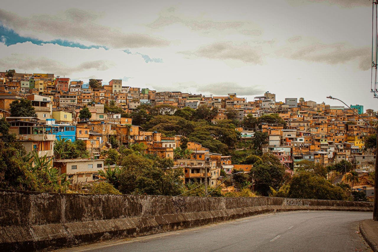 A imagem mostra a vista do Aglomerado da Serra, visto da descida da Av. do Cardoso. A paisagem mostra a pista em primeiro plano e ao fundo as casas do aglomerado.