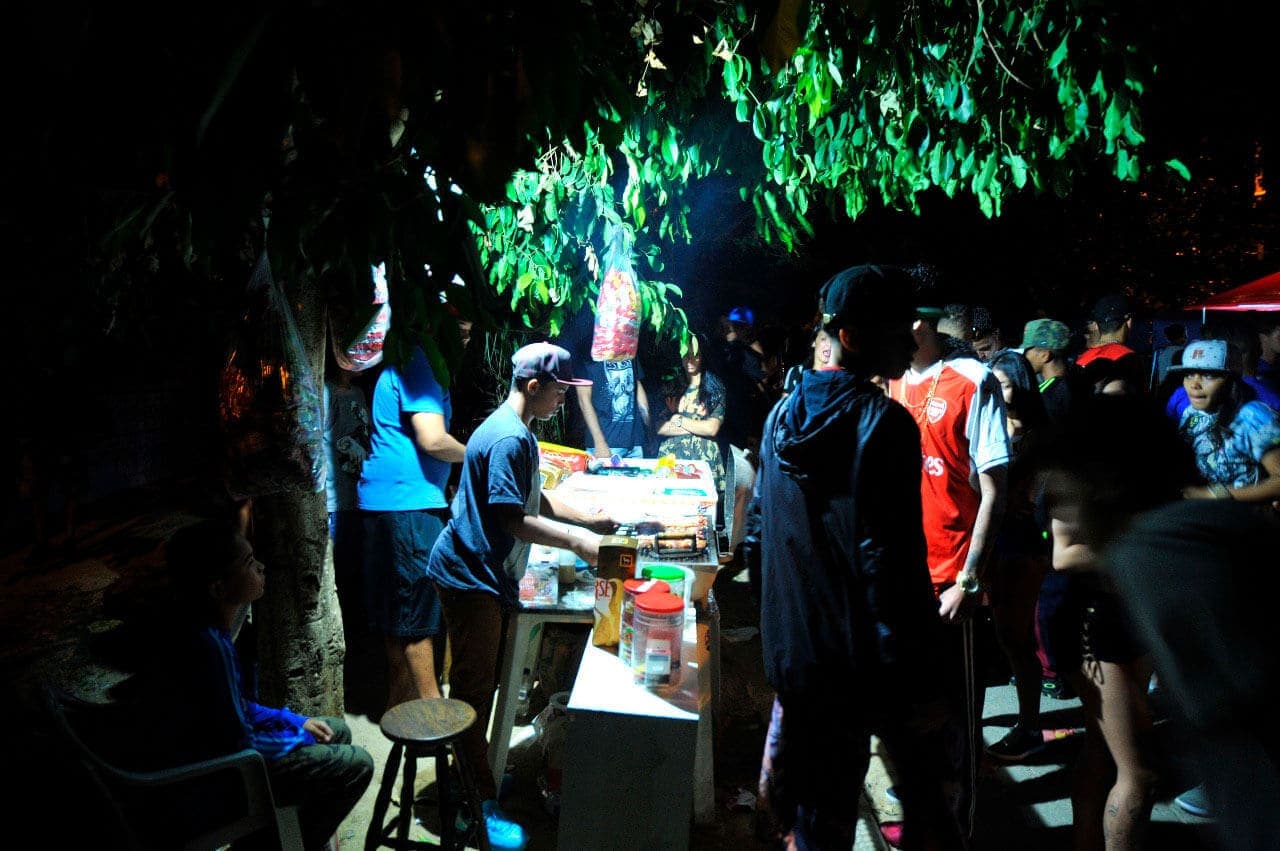 A imagem mostra uma barraquinha com venda de comida no Baile da Serra. É uma foto noturna, onde é possivel ver as pessoas que estão participando do evento e com uma iluminação mais forte o local onde acontecem as vendas. Um garoto de boné está fazendo o atendimento.