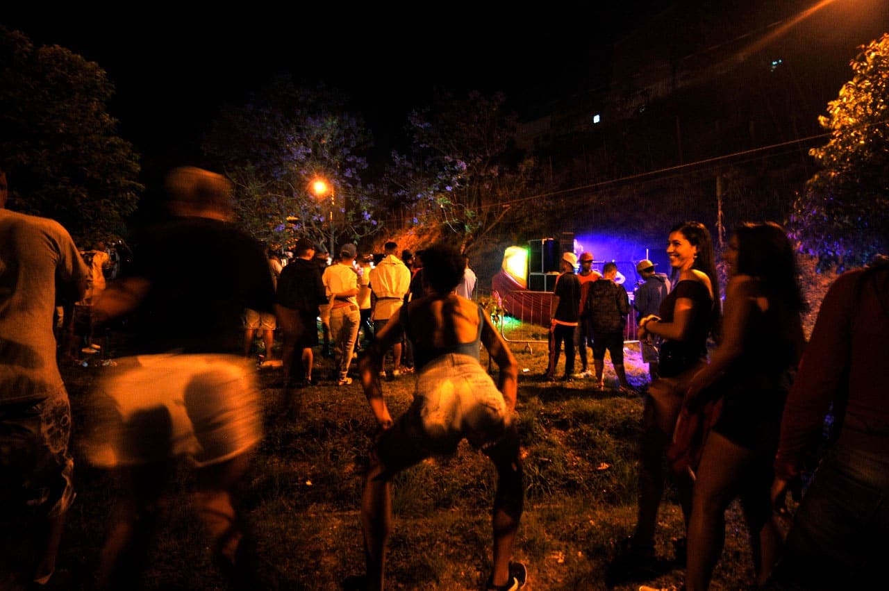 A imagem mostra uma edição noturna do Baile da Serra. É possível ver em primeiro plano pessoas de costas, com a mão no joelho dançando funk, sobre um gramado. Ao fundo é possível visualizar mais pessoas que estão presentes no evento.