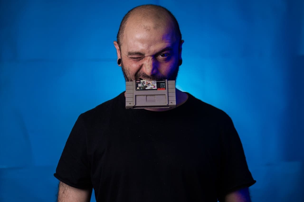 Flávio é um homem branco, com poucos cabelos. Ele posa de blusa preta em frente a um fundo azul. Enquanto olha para a câmera, morde um cartucho antigo de videogame. Além disso, ele possui alargador nas orelhas.