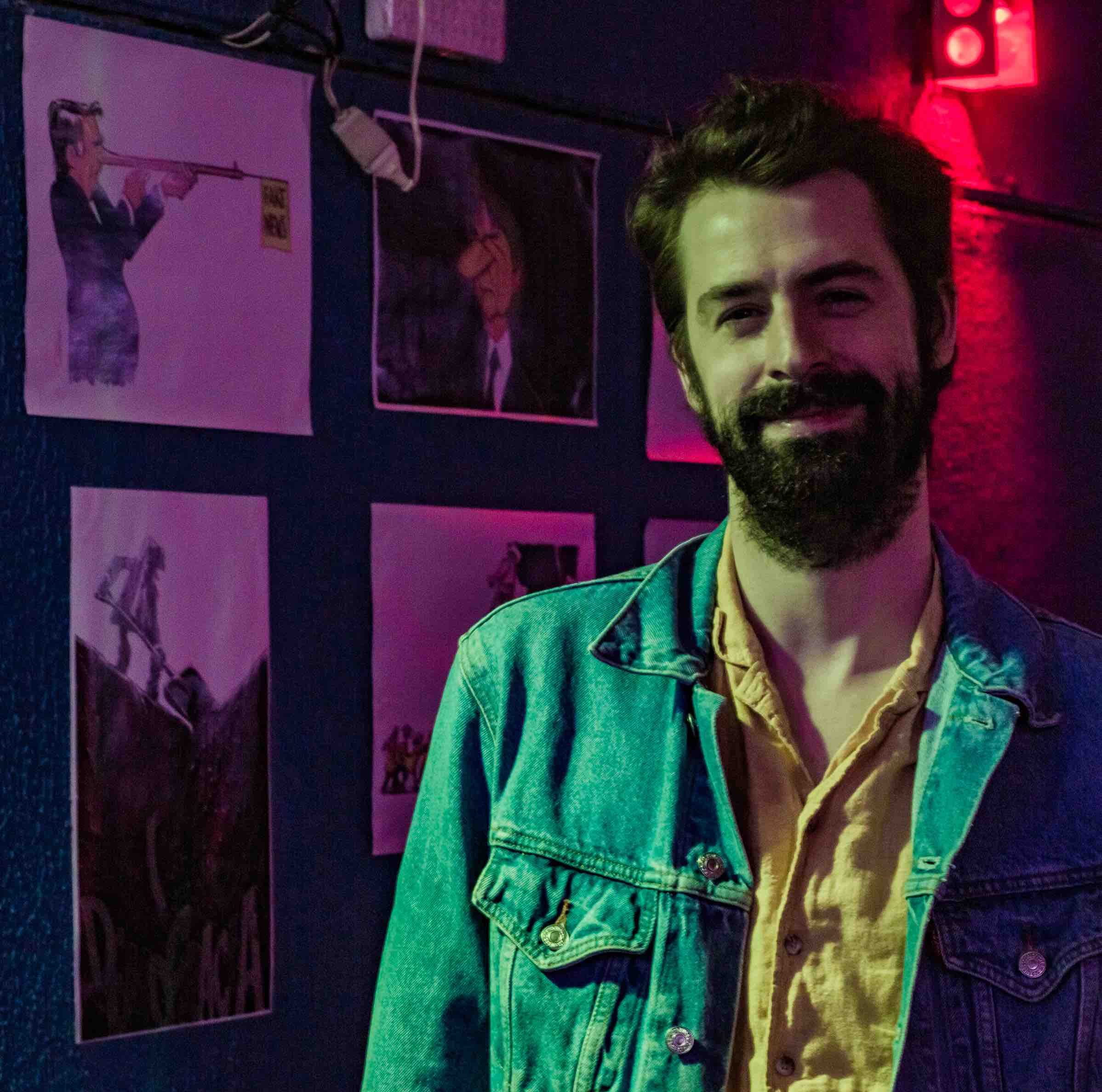 A foto mostra Leo Bryan, um homem branco com barba e cabelos escuros, vestindo uma camisa amarela e uma camisa jeans por cima dela.