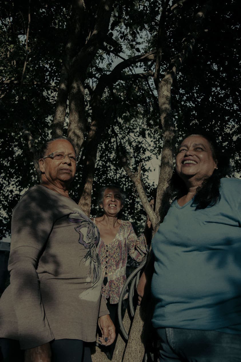 A foto mostra três mulheres sorridentes do Quilombo Família Souza. Uma está mais ao fundo, no meio, e usa um vestido estampado. A que está a direita usa os cabelos pretos soltos e uma blusa azul claro. A que está à esquerda é a única que olha para a câmera. Ela usa óculos e usa uma blusa cinza com uma estampa no meio. Ao redor das mulheres estão galhos de árvores.