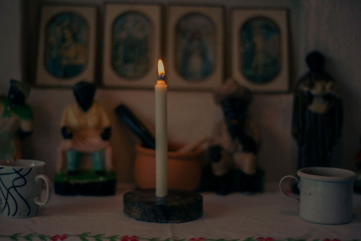 A foto mostra um detalhe do altar que fica em um dos cômodos do quilombo. Ao centro, uma vela branca acesa apoiada em um píres preto. Dos lados direito e esquerdo, duas xícaras. Atrás, imagens de preto-velho. Presos na parede, quadros pequenos com imagens de santas.
