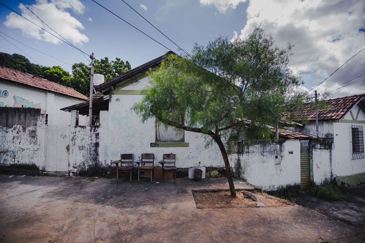 A foto mostra uma das casas da Cidade Ozanam de frente, com 3 cadeiras antigas de madeira vazias em frente à casa. Em primeiro plano, uma pequena árvore em frente a ela. O céu está azulado com algumas nuvens. A casa é branca e tem a pintura desgastada. Alguns detalhes na empena e a janela estão pintados de verde e o telhado é de cerâmica em duas águas.