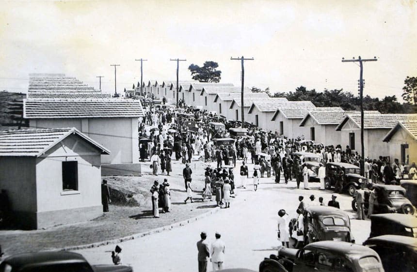 A foto, em preto e branco, mostra uma rua de longe e as casas da Cidade Ozanam enfileiradas em perspectiva. No meio da rua, há uma grande aglomeração de pessoas, ali presentes para a festa de inauguração da Cidade Ozanam. Também se vê alguns carros típicos do final da década de 1930.