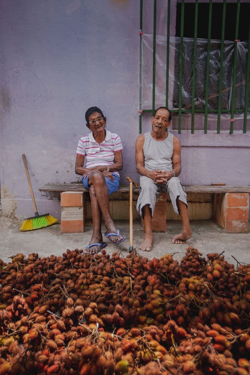 A foto mostra uma senhora de óculos, blusa listrada de branco e vermelho, bermuda azul e chinelo azul. Ao seu lado, um senhor de regata e calça cinza. Os dois estão sentados em um banco formado por uma tábua de madeira apoiada em três pilares de tijolos. Entre os dois, uma bengala. Ao lado da senhora, uma vassoura de cerdas veres e amarelas. A fundo, uma parede lilás e as grades de uma janela verde. No chão de cimento, galhos de frutos de urucum.