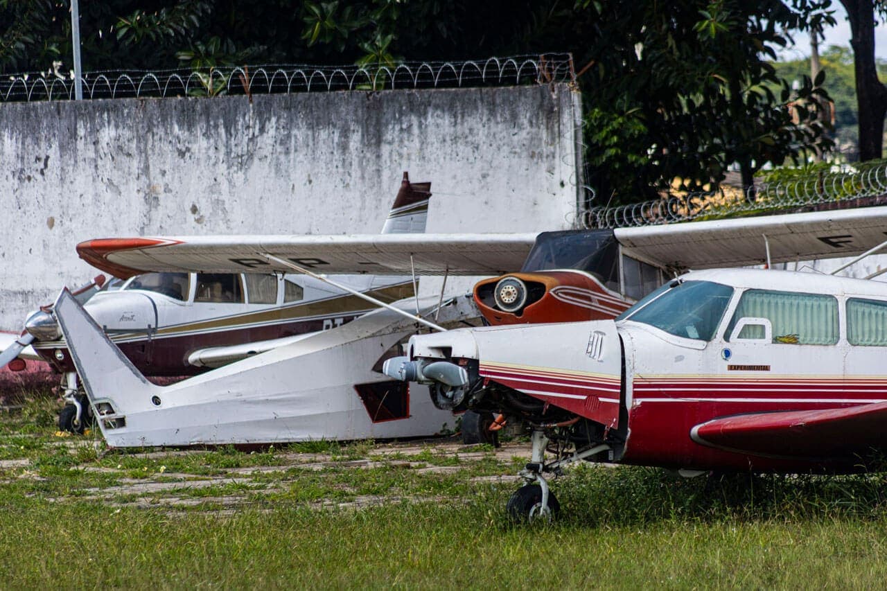 A imagem traz quatro aeronaves de modelo mais antigo e porte pequeno aparecem estacionadas, lado a lado. Uma delas é vermelha e branca e parte da lataria do bico onde fica a hélice não está na aeronave, outra é marrom e branca, outra é totalmente branca e outra é branca com faixas vermelho e marrom. Apenas uma parte de cada uma das aeronaves aparece no enquadramento.