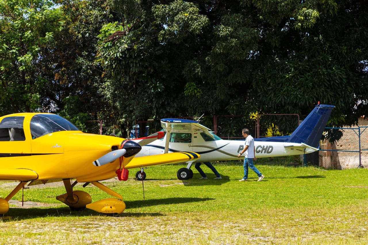 A imagem mostra duas aeronaves de pequeno porte paradas na grama. A aeronave à esquerda é amarela e só conseguimos ver sua parte dianteira. A aeronave que está ao centro, atrás da amarela, é branca com detalhes em azul e marrom claro. Um homem caminha próximo a aeronave branca. Ele está de tênis, calça jeans e blusa social branca. Atrás das aeronaves, uma cerca de arame e árvores que cobrem a parte de cima da foto.