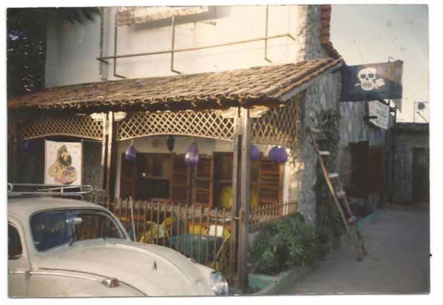A foto mostra uma casa antiga com uma varanda onde estao diversas mesas e cadeiras, além de lustres roxos. Em frente a esta casa, tem um fusca branco que aparece só pela metade. Nessa casa funcionava o antigo Bar Calabouço.