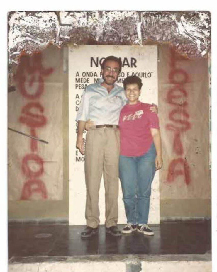 A foto antiga mostra Sônia Soares e Patrus Ananias bem no centro. Patrus é um homem branco, com cabelos e barba escuros. Ele usa óculos, uma camisa social azul clara e calça social bege. Sônia é uma mulher branca, de cabelos curtos e escuros. Ela vez uma camisa vermelha, calça jeans e tênis. Eles posam em frente a uma parede pixada com as palavras “música” e “poesia”.