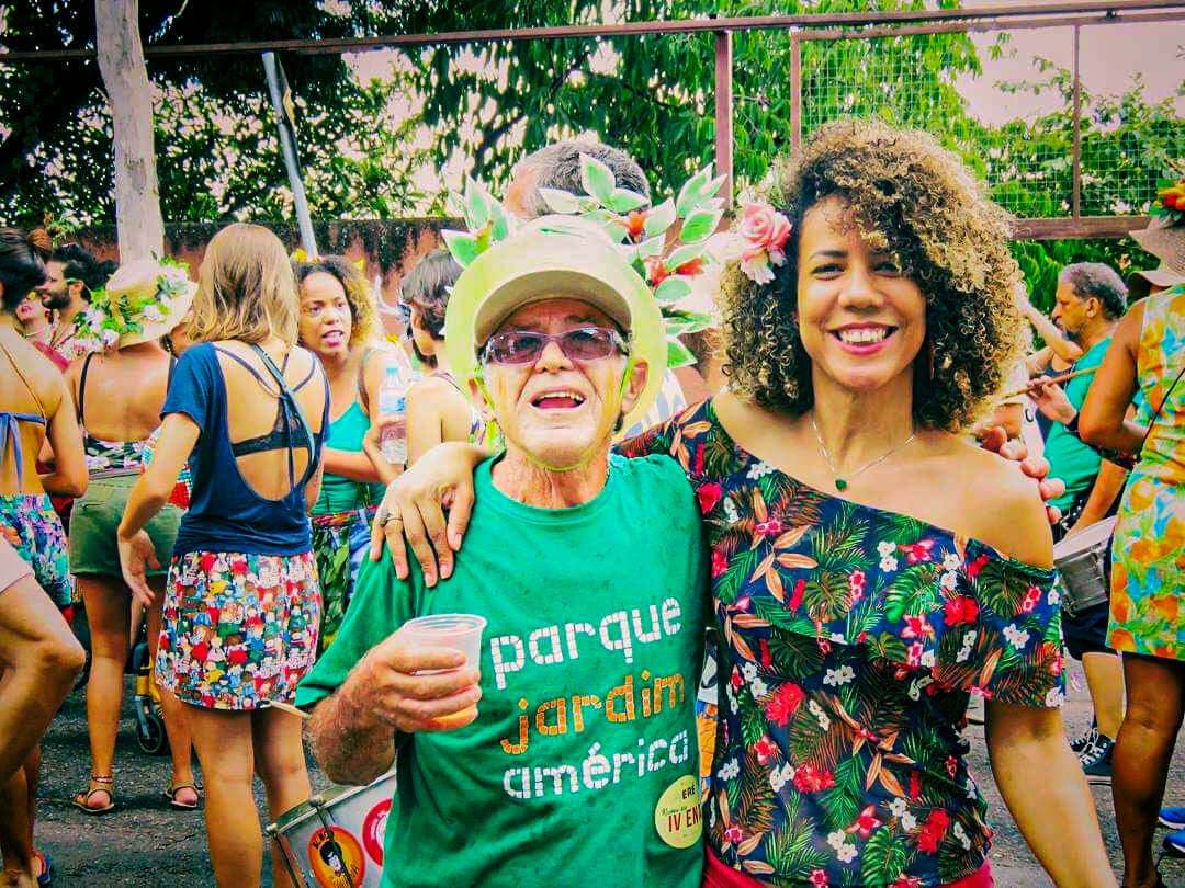 A imagem mostra um senhor de pele clara, cabelos grisalhos, óculos vestindo uma camisa verde escrito “Parque Jardim America”. Ele carrega um copo em uma mão e abraça uma mulher negra de cabelos enrolados e blusa florida. Os dois sorriem para a foto. Ao fundo, diversas pessoas de blusas floridas e algumas pessoas carregando instrumentos de percussão.