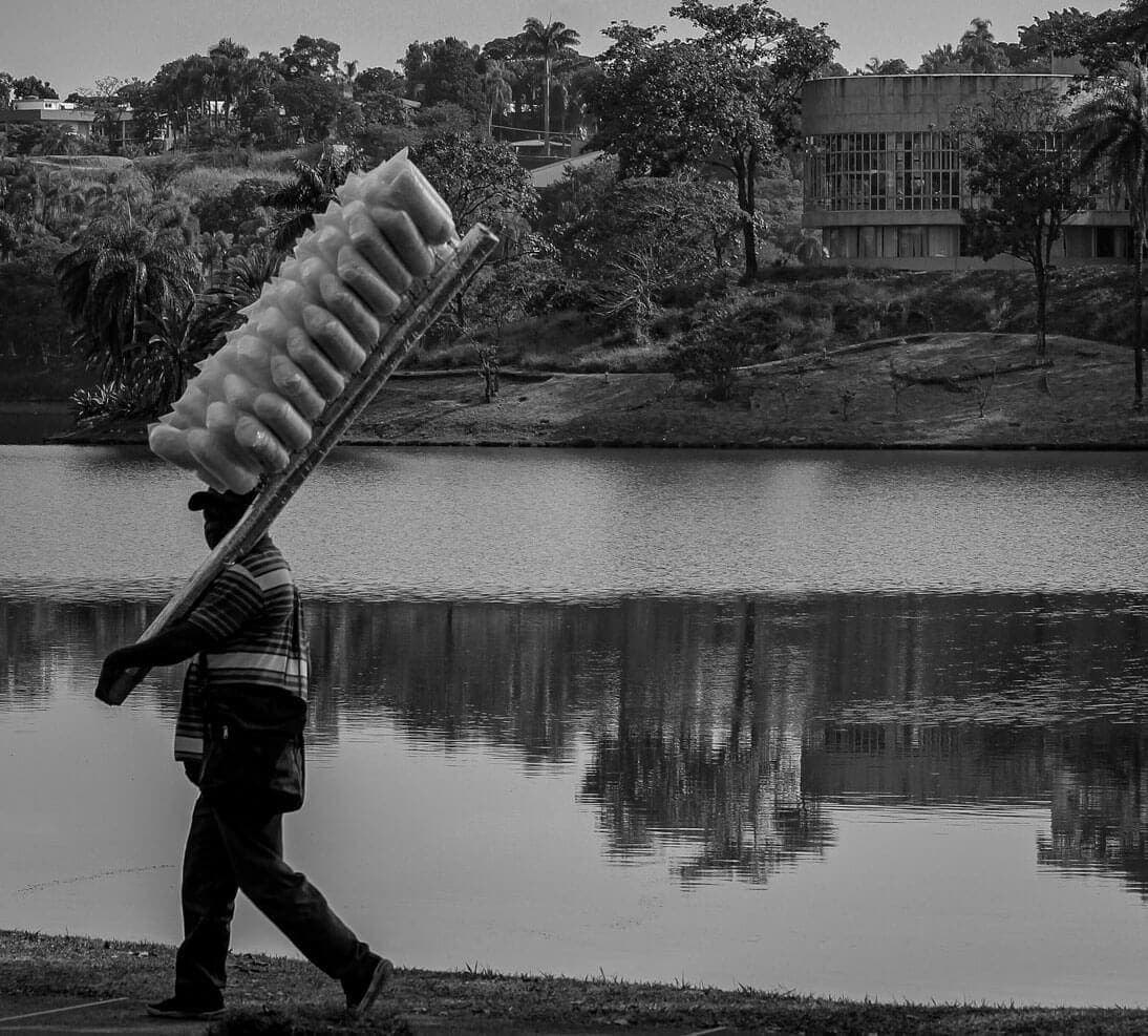 A imagem mostra uma paisagem com uma lagoa abaixo e uma construção moderna do lado direito. O restante são árvores e vegetação ao redor. Do lado esquerdo da foto, aparece um vendedor de algodão doce com os sacos pendurados em um pau que ele ergue com as mãos.