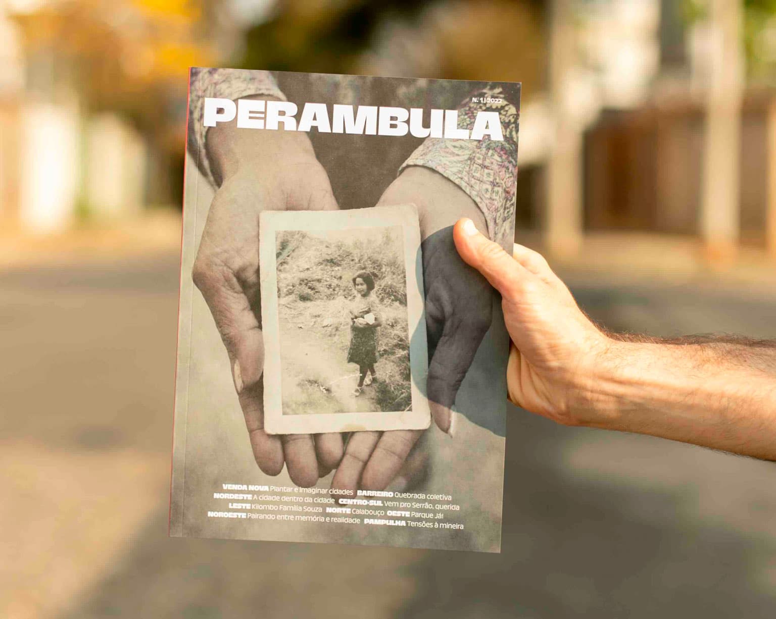 A foto mostra o braço de uma pessoa branca segurando a revista Perambula. Ao fundo, com aspecto borrado, é possível perceber uma pasaigem de rua com calçamento de pedras. Na capa da revista há uma fotografia em que um par de mãos de uma mulher negra segura um retrato antigo de sua avó quando criança. 