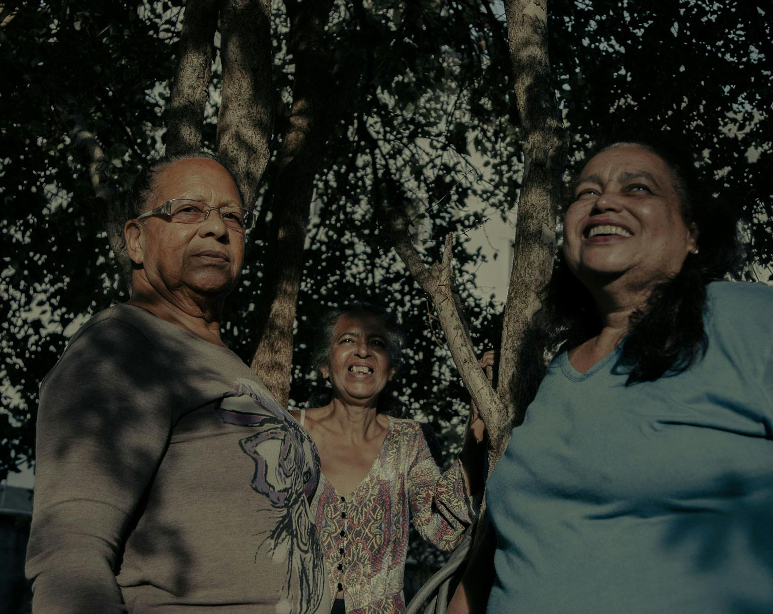 A foto mostra três mulheres sorridentes do Quilombo Família Souza. Uma está mais ao fundo, no meio, e usa um vestido estampado. A que está a direita usa os cabelos pretos soltos e uma blusa azul claro. A que está à esquerda é a única que olha para a câmera. Ela usa óculos e usa uma blusa cinza com uma estampa no meio. Ao redor das mulheres estão galhos de árvores.