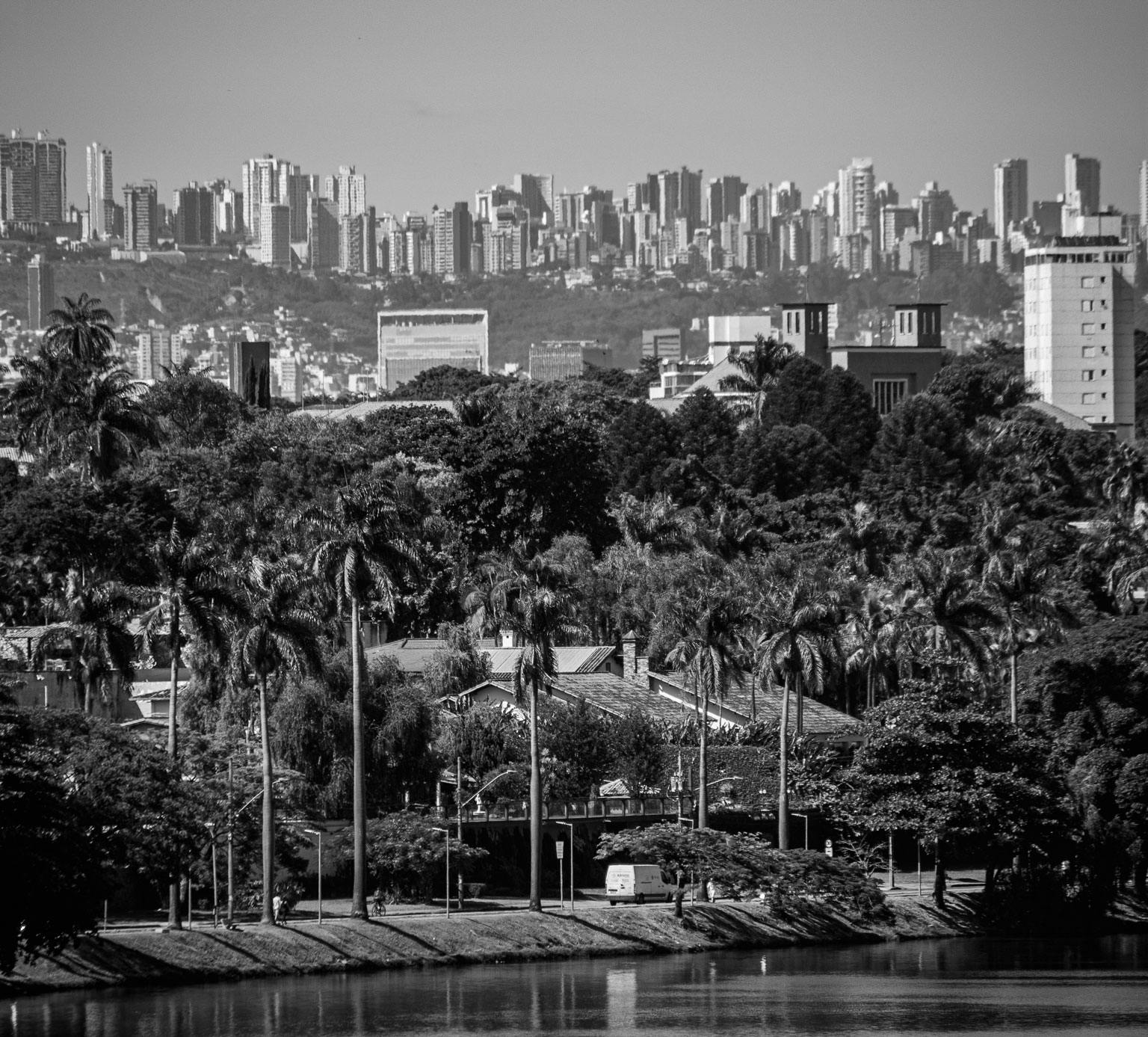 A imagem é uma paisagem de Belo Horizonte vista da Lagoa da Pampulha. No fundo é possível ver vários prédios, pequenos. Mais perto, vemos um monte de árvores que rodeiam a lagoa.