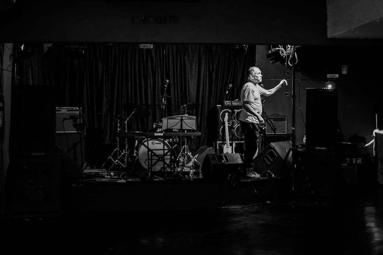 A foto, em preto e branco, mostra Edmundo em cima de um palco com pouca iluminação, arrumando os equipamentos antes de um show na Casa Matriz. Edmundo é um homem branco, com cabelos grisalhos na altura do ombro. Ele veste camisa de manga curta e manga comprida.