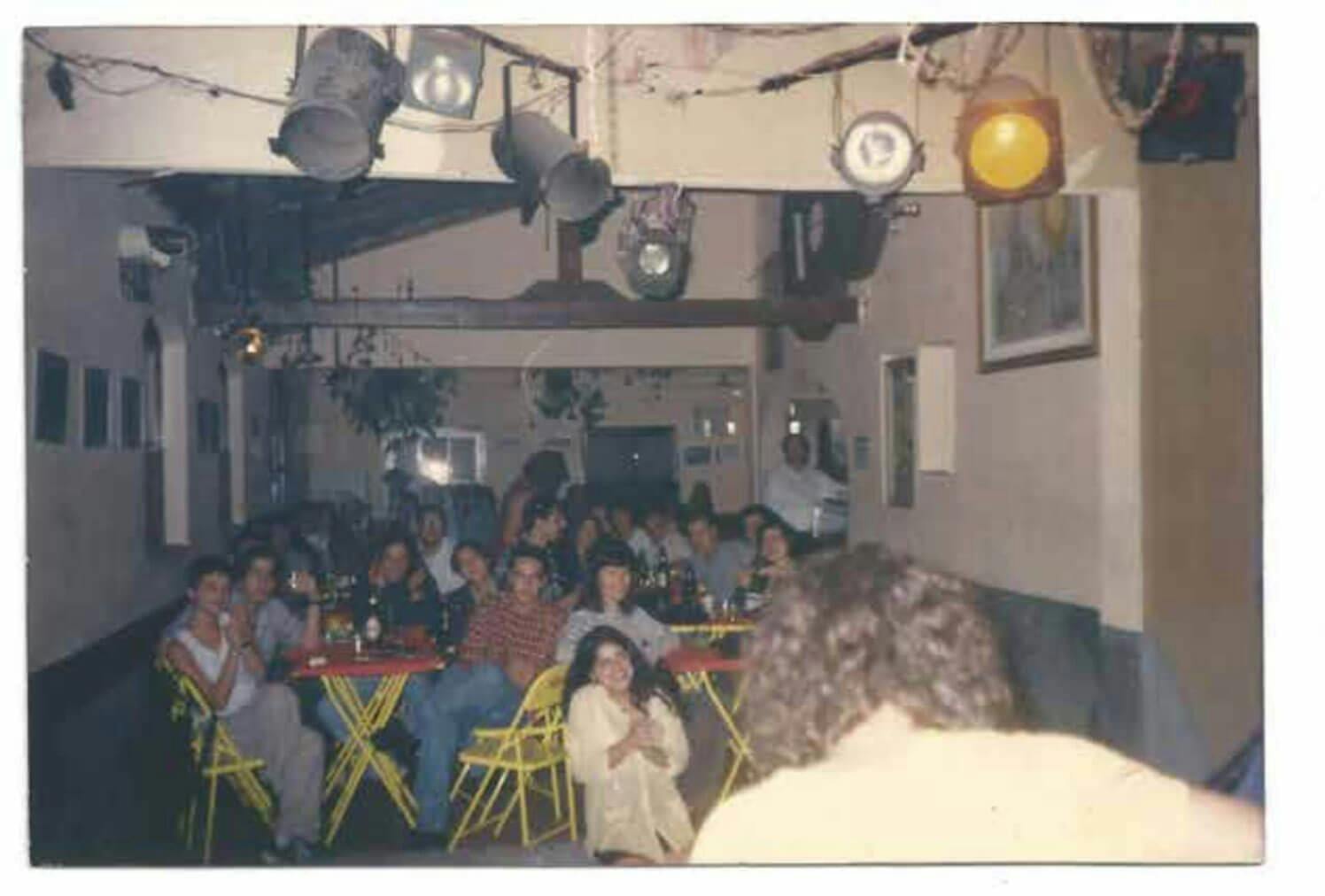 A foto mostra o interior do Bar Calabouço, do ponto de vista do palco. Em primeiro plano, as costas do músico que se apresenta. Ao fundo, diversas pessoas sentadas em mesas e cadeiras de ferro amarelas.