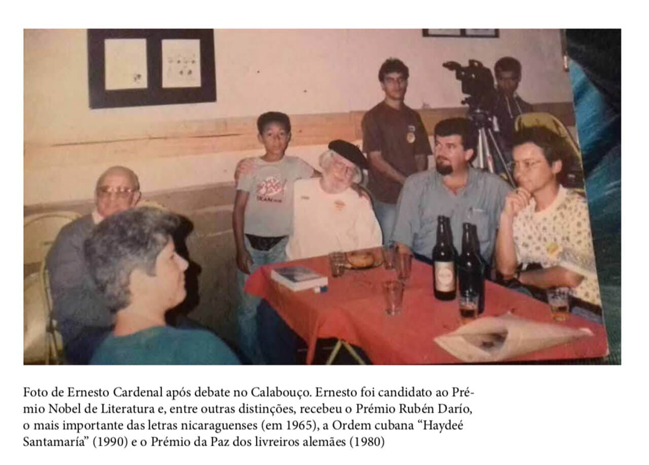 A foto mostra pessoas reunidas em voz de Ernesto Cadernal, um homem branco com cabelos e barba brancas, vestindo um camisa branca e um chapéu preto. Ele está sentado de frente a uma mesa com toalha vermelha e garrafas de cerveja em cima.